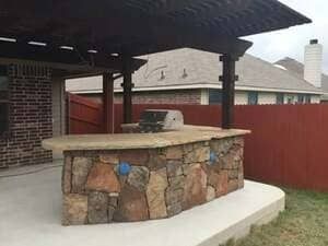 An outdoor stone kitchen counter with a grill, situated under a wooden patio cover on a concrete slab in a backyard.