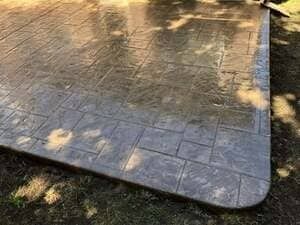 A close-up view of a gray stamped concrete patio with a rectangular stone pattern border and dappled sunlight.