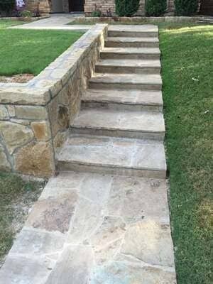 A stone walkway leading up a set of stone steps beside a low stone retaining wall outdoors.