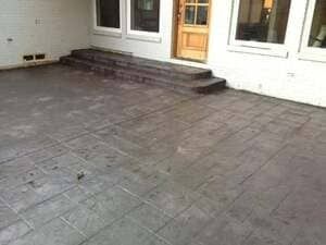 A gray, rectangular-patterned concrete patio leading to wooden steps and a doorway at the back of a white building.