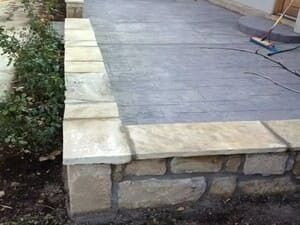 A patio with a stamped concrete surface, bordered by a low, natural stone retaining wall topped with light stone caps.