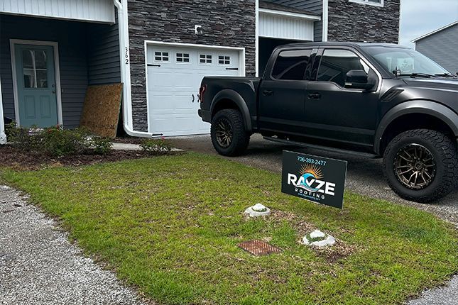 A dark gray pickup truck parked in front of a house, with a Rayze Roofing sign on the front lawn.