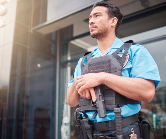 An armed security guard in a police uniform stands outside a building, attentively monitoring the area.