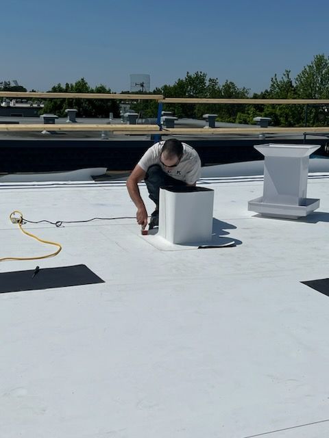 Un homme sur un toit blanc scellant un évent, ciel bleu au-dessus.