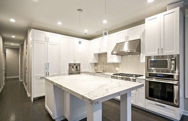 Bright white kitchen with wooden floors, island counter, stove, sink, and sunlit windows with blinds.