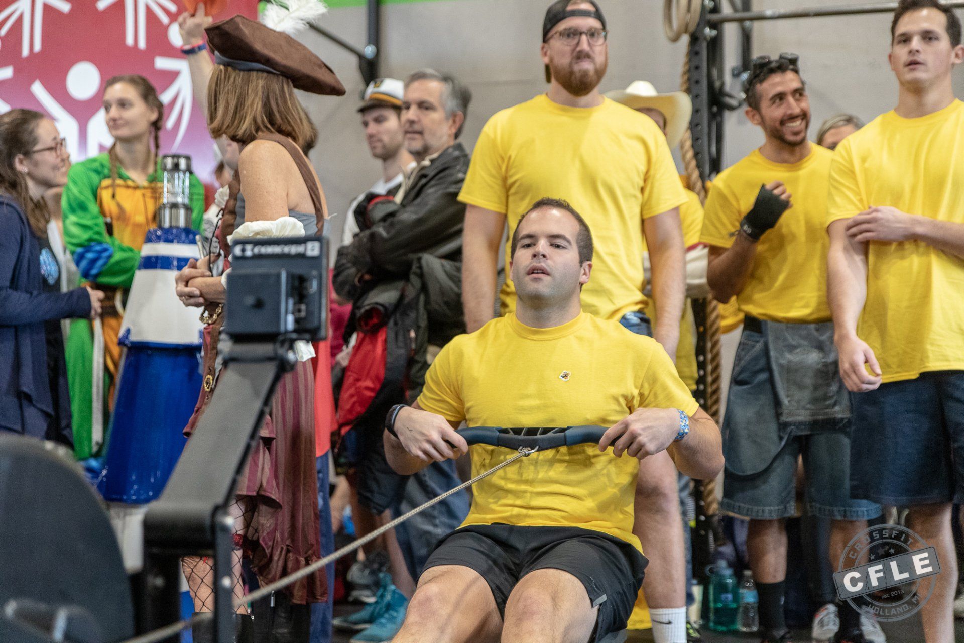A group of people are standing around a man rowing a rowing machine.