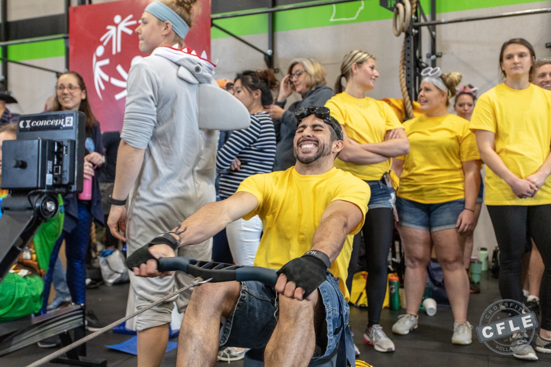 A man is riding a rowing machine in a gym while a group of people watch.