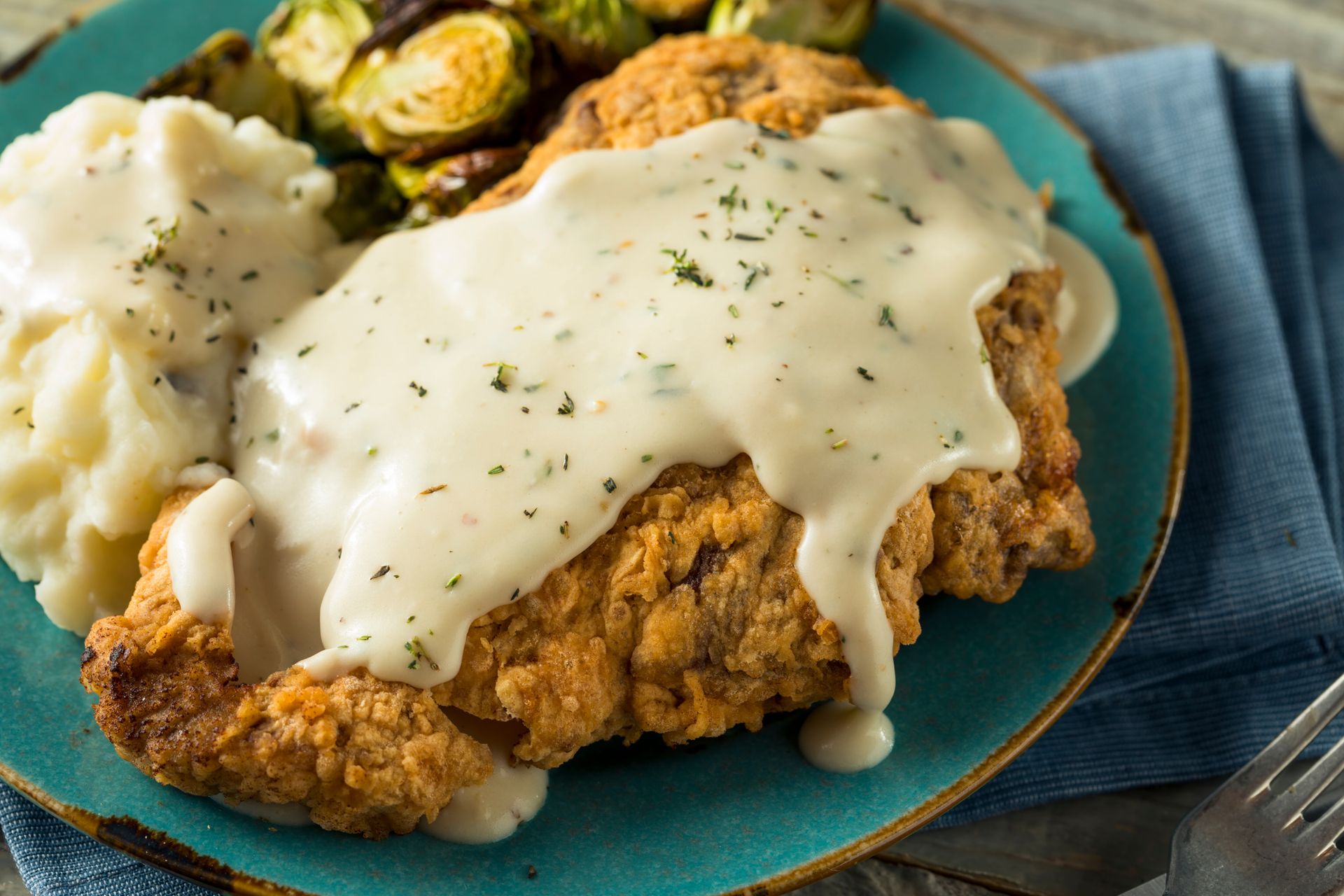 Fried chicken cutlet with white gravy, mashed potatoes, and Brussels sprouts on a blue plate