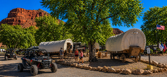 Covered wagon on a dirt path near trees and red rock cliffs in a warm, painted landscape