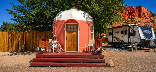 Round yurt cabin before red cliff, with small porch, trees, and a campfire in a desert landscape