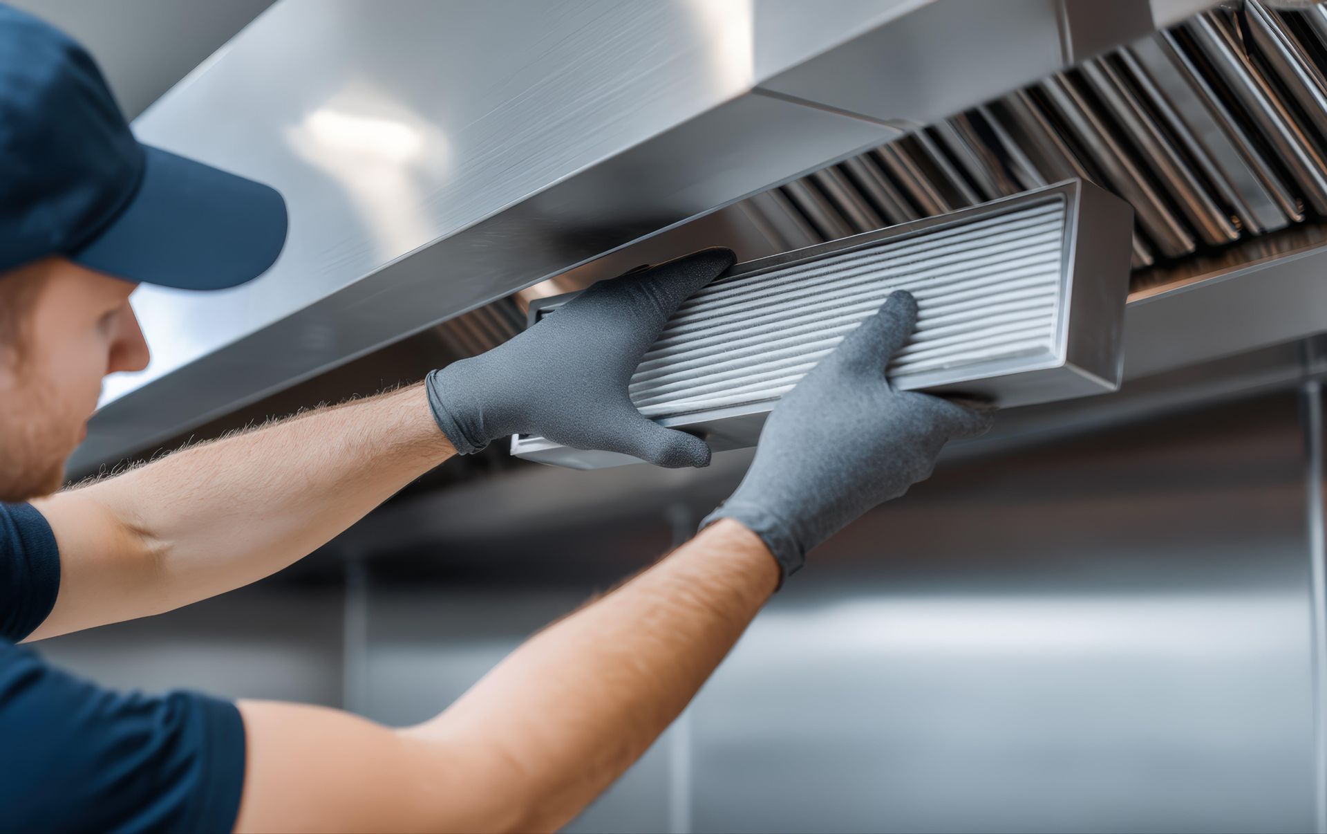 A technician's gloved hands, removing a kitchen hood filter for maintenance and inspection.