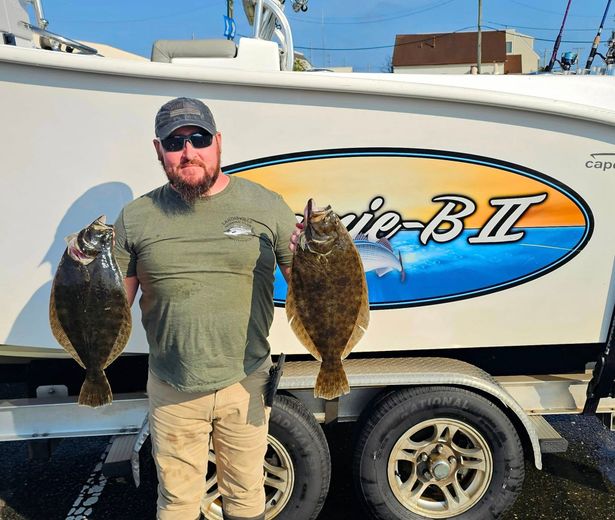 A man is holding two fish in front of a boat that says cape-b-ii