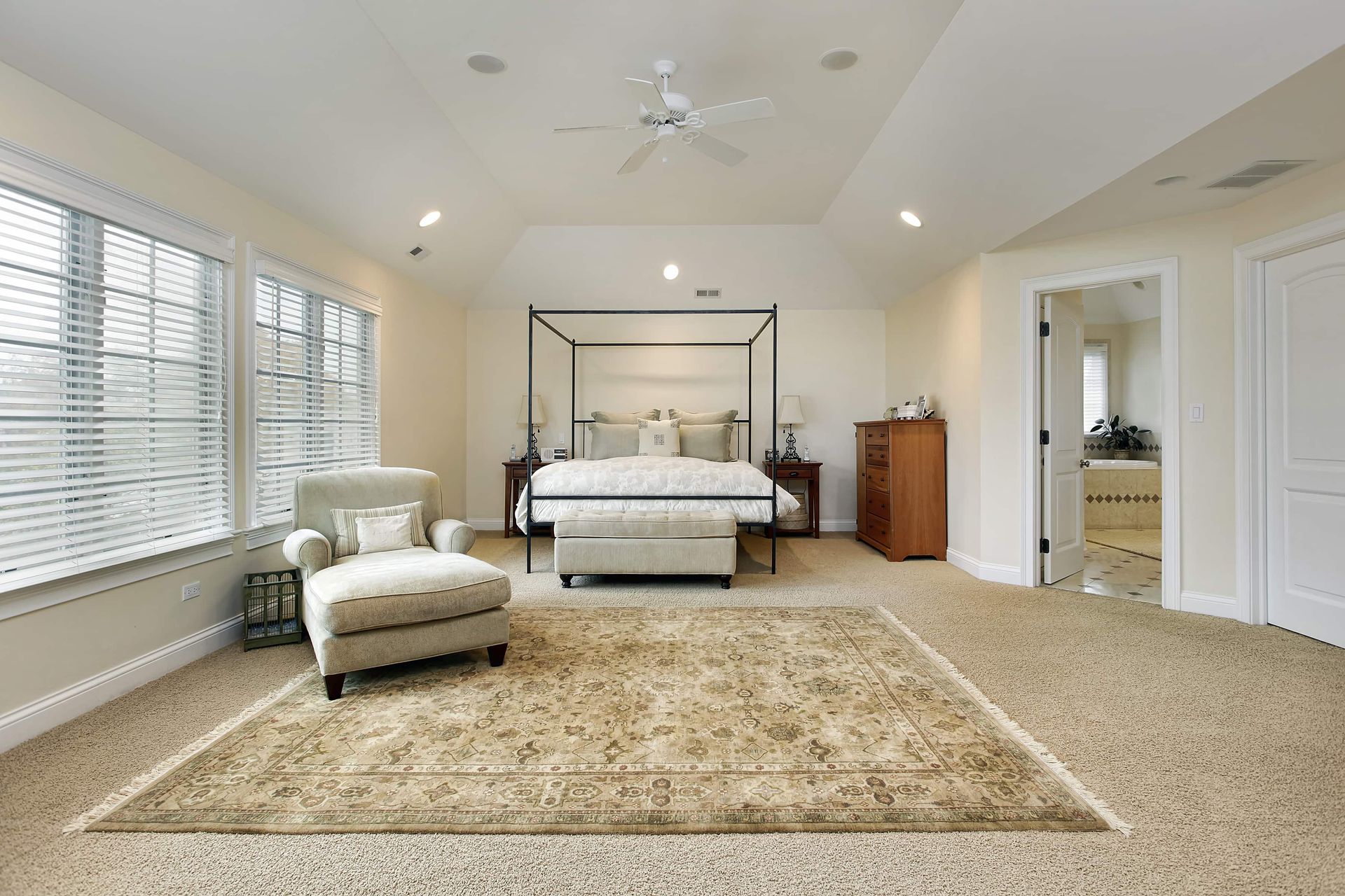 Spacious beige bedroom with canopy bed, armchair, oriental rug, and a doorway to another room.