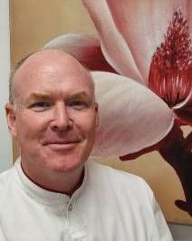 A Man in a White Shirt is Smiling in Front of a Painting of a Flower — Goonellabah Denture Clinic in Goonellabah, NSW