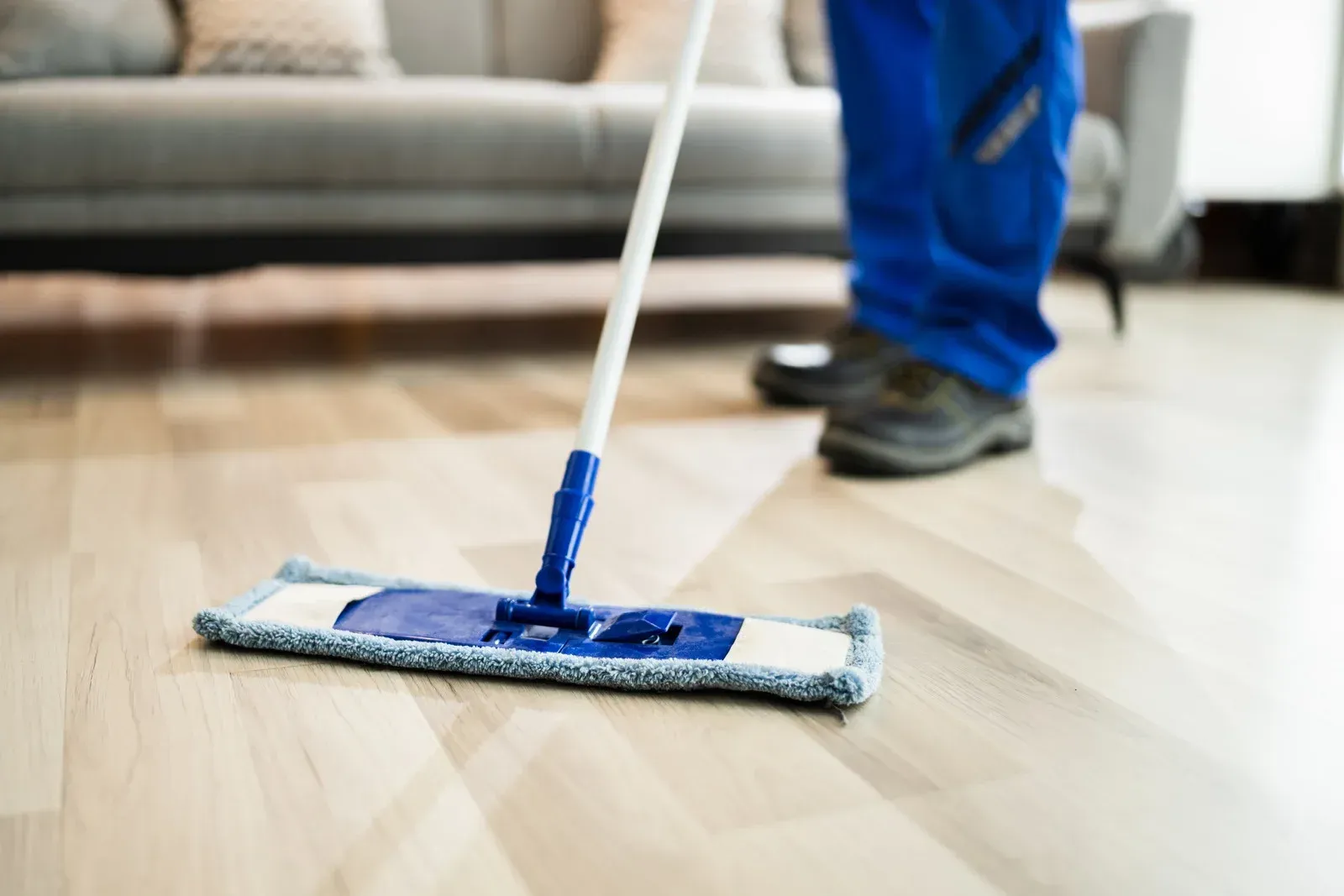 Person mopping a hardwood floor in a living room, wearing blue work clothes.