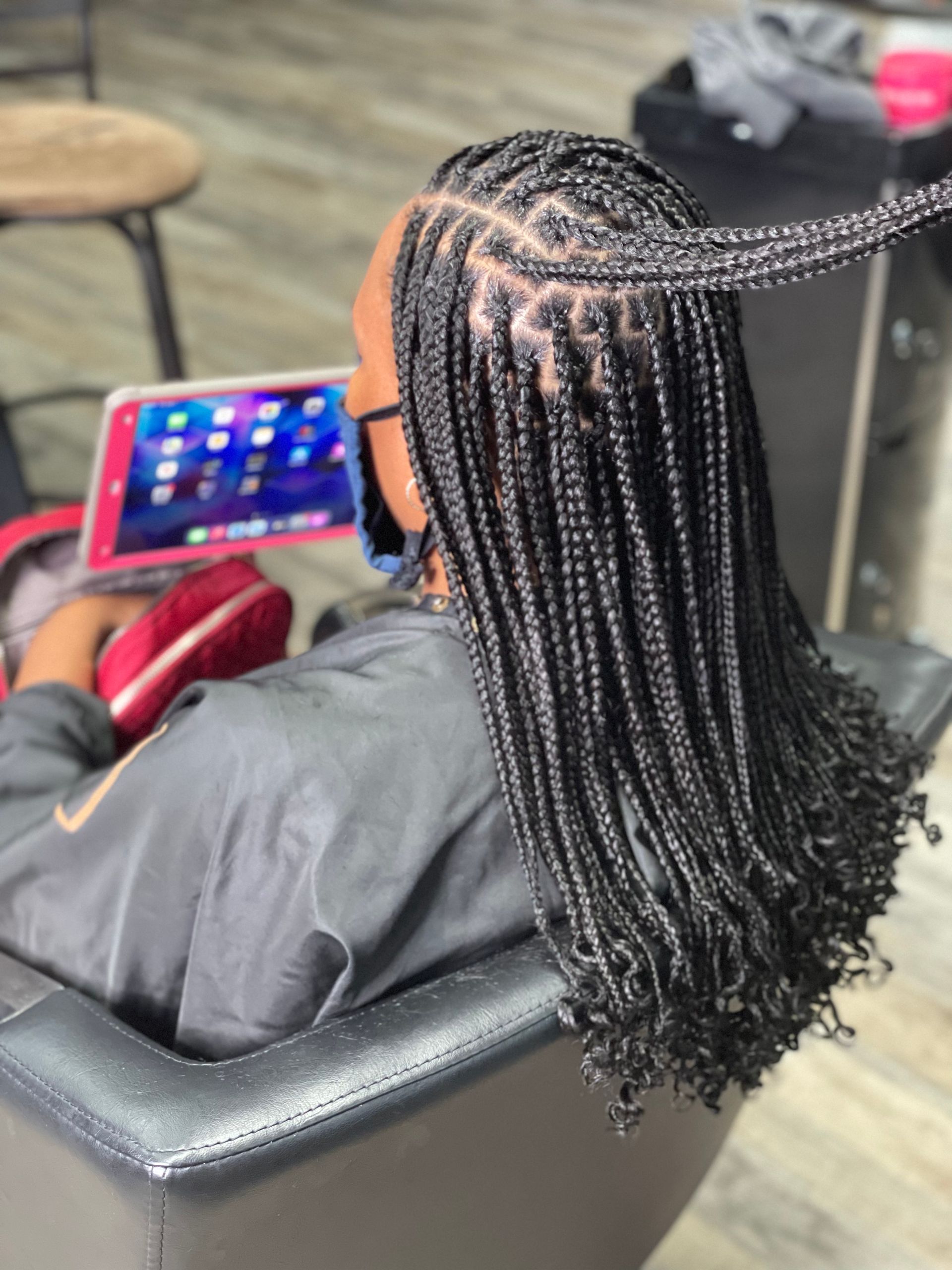 Person with box braids in a salon, looking at a tablet. Braids are long, black, and curly at the ends.
