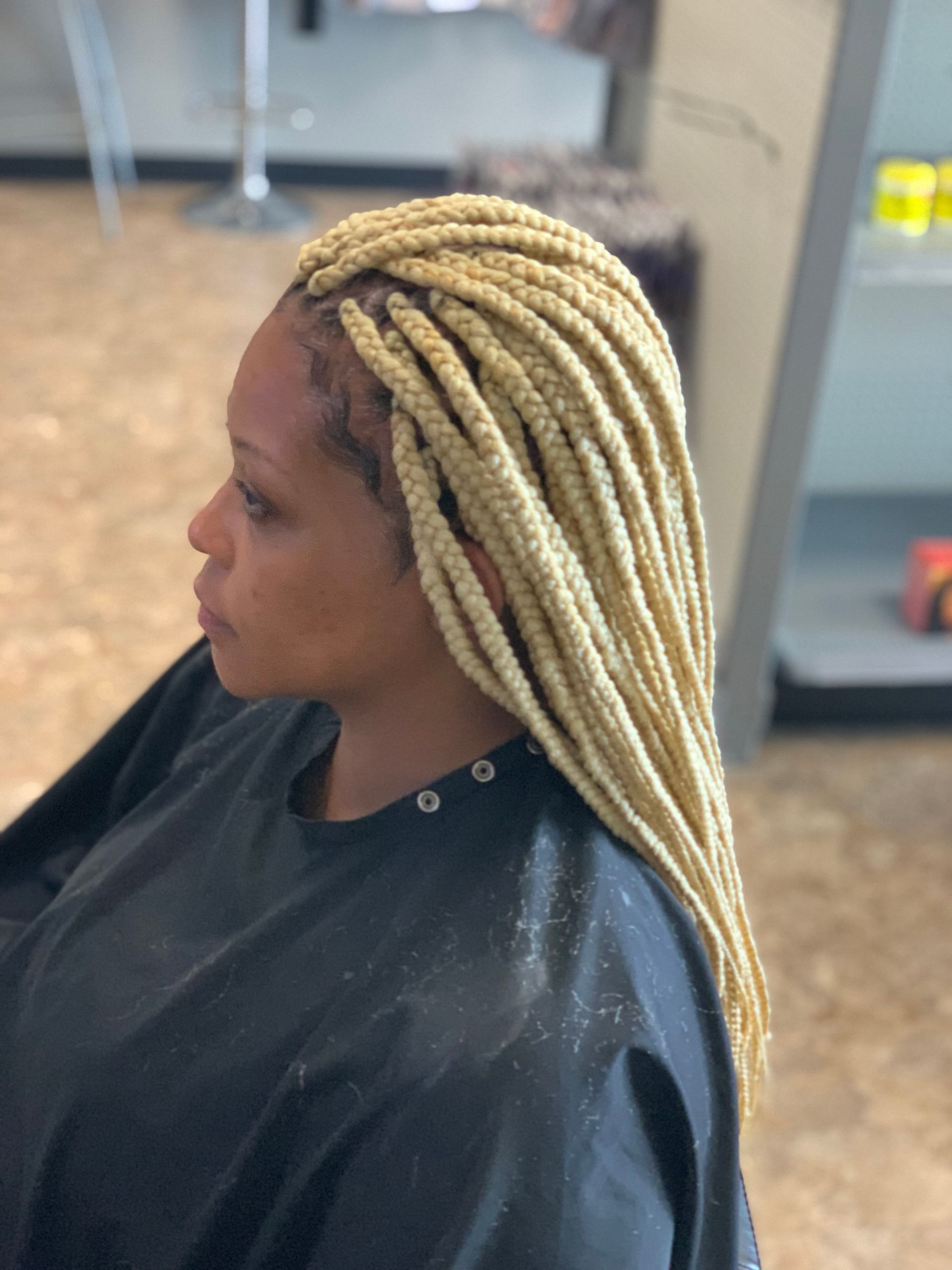 Woman with blonde box braids in a salon setting. She wears a black cape and looks to her left.