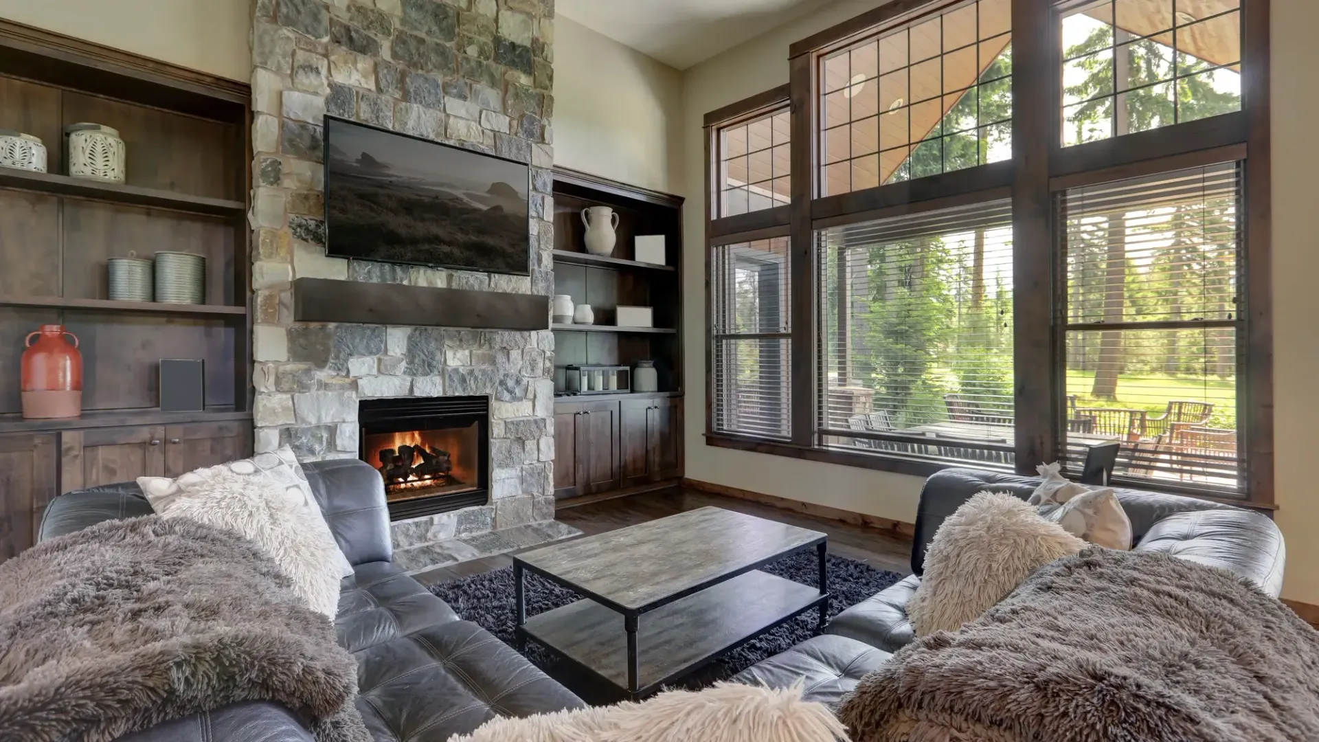 Cozy living room with fireplace, stone wall, large windows overlooking trees, and plush sofa with blankets.