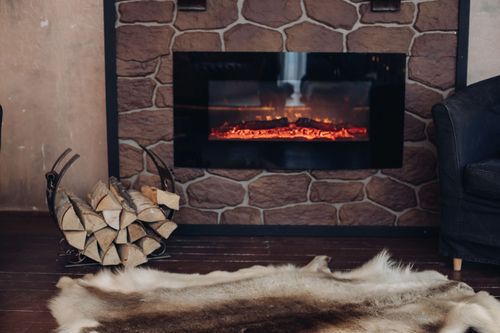 Fireplace with a stone surround and glowing flames, next to firewood basket and rug.
