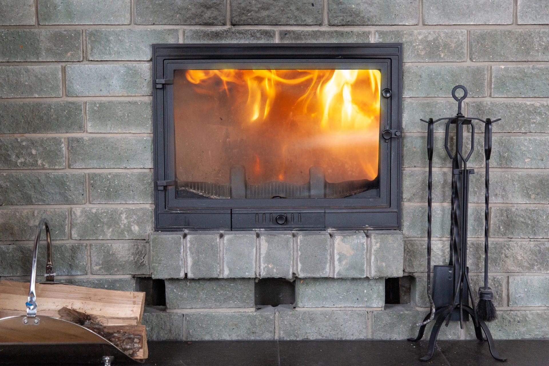 Fireplace with flames burning, built into gray brick wall. Fireplace tools on the right.