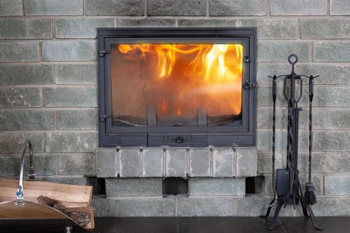 Fireplace with flames burning, built into gray brick wall. Fireplace tools on the right.