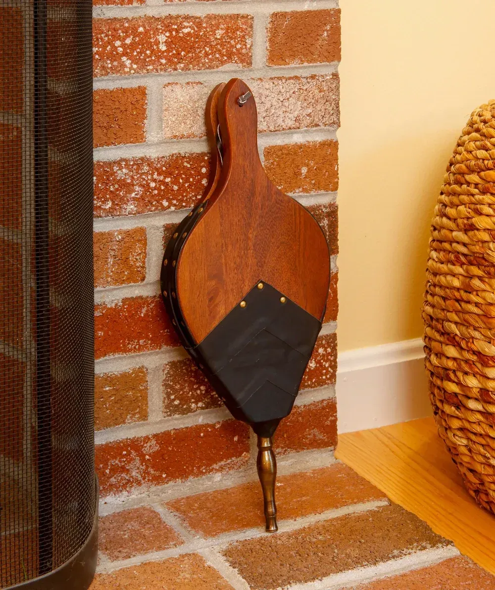 Fireplace bellows made of wood and leather hanging on a brick wall next to a woven basket.