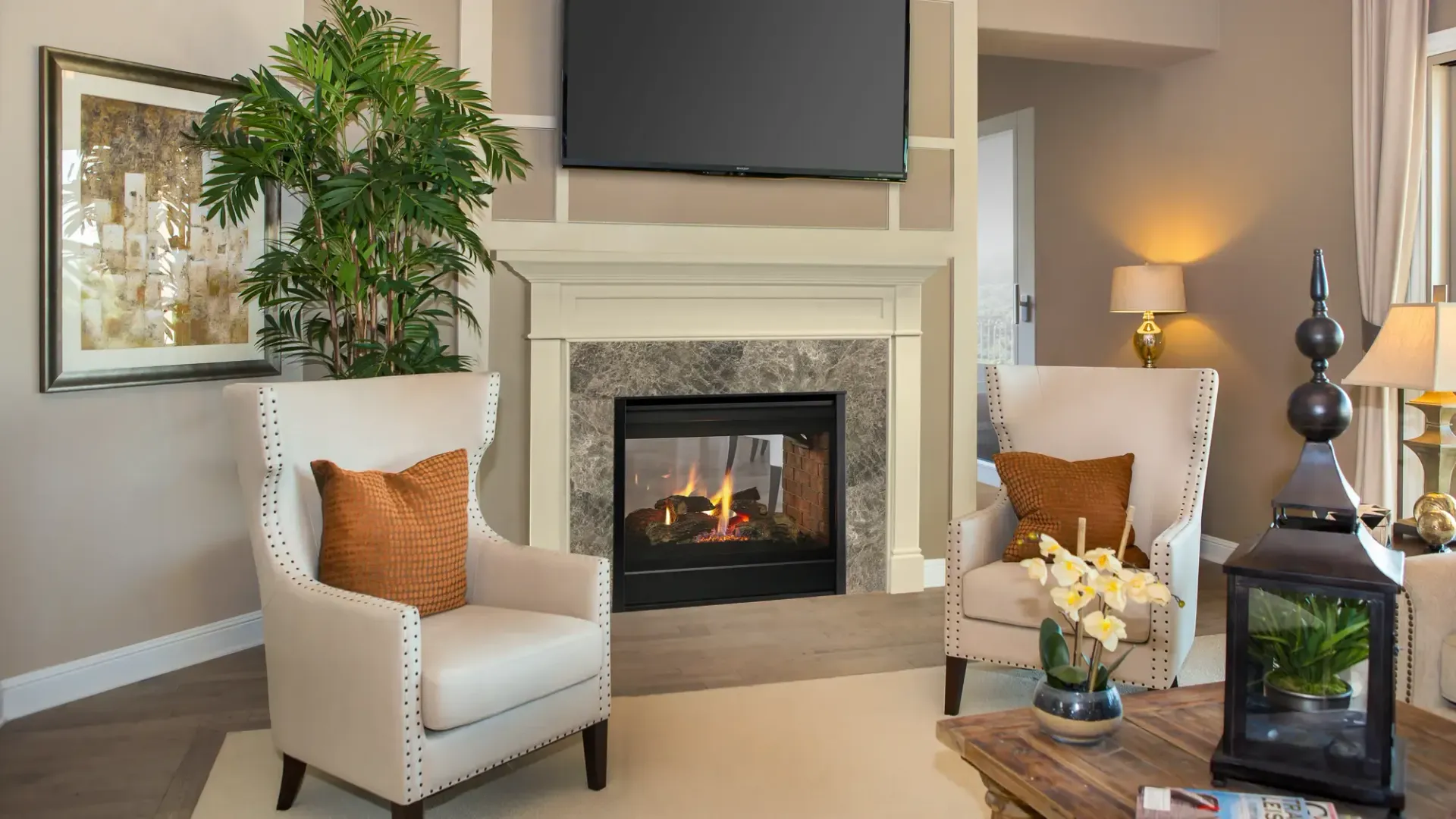 Living room with fireplace, armchairs, and large TV. Neutral tones, cozy setting.