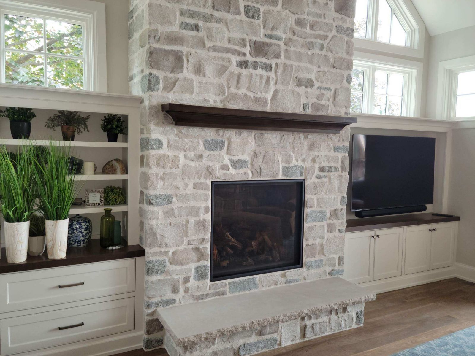Stone fireplace with dark mantel and built-in cabinets. TV on right, plants and decor on left.