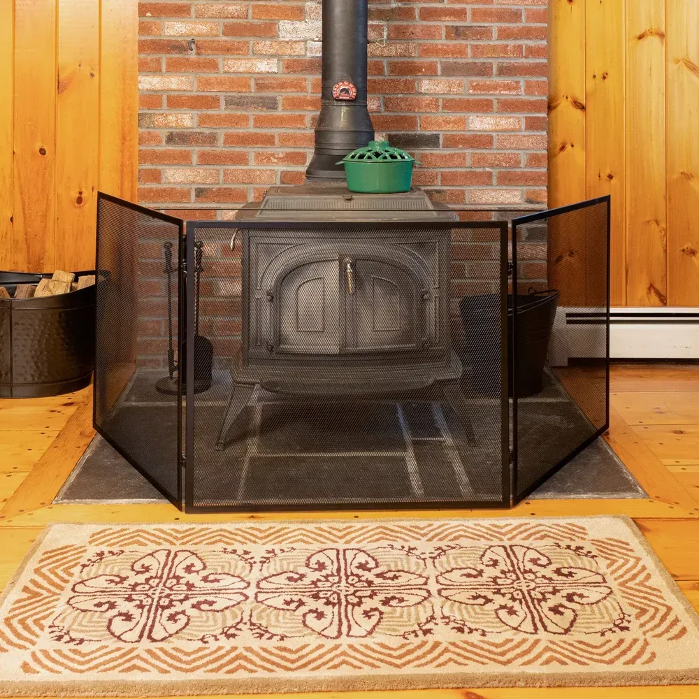 Black fireplace screen in front of a wood-burning stove. Red brick chimney, wood paneling, patterned rug.