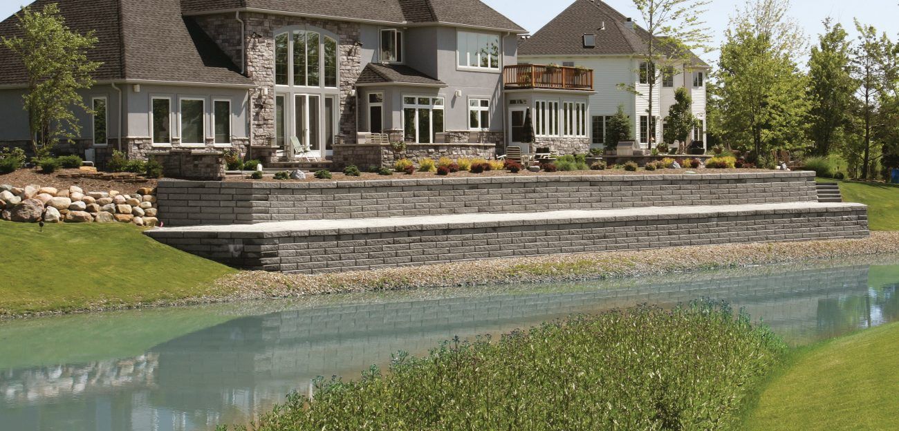 A large, gray house with a stone facade next to a retaining wall and a lake.