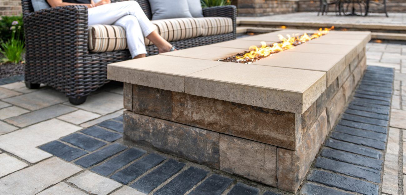 Outdoor fire pit with person on a wicker couch. Stone pavers and flames are visible.