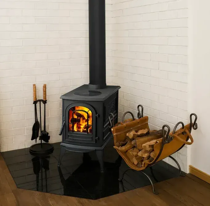 Wood-burning stove with burning flames, surrounded by tools and firewood, set against a white brick wall.
