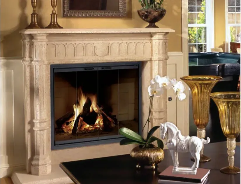 Fireplace with burning logs, ornate tan stone mantel, decor on a coffee table, and vases.