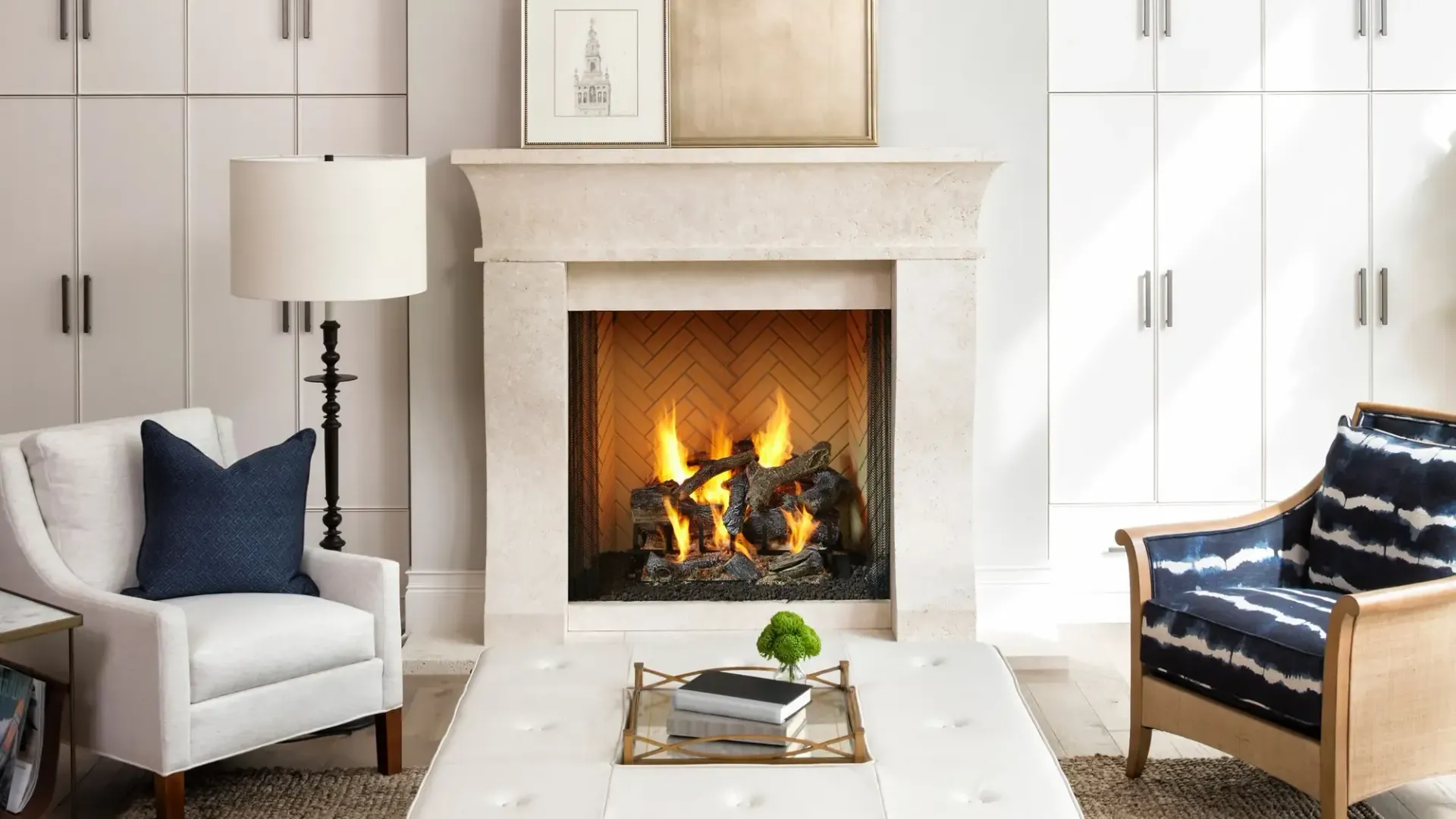 Living room with a lit fireplace, white walls, chairs, and a white ottoman.