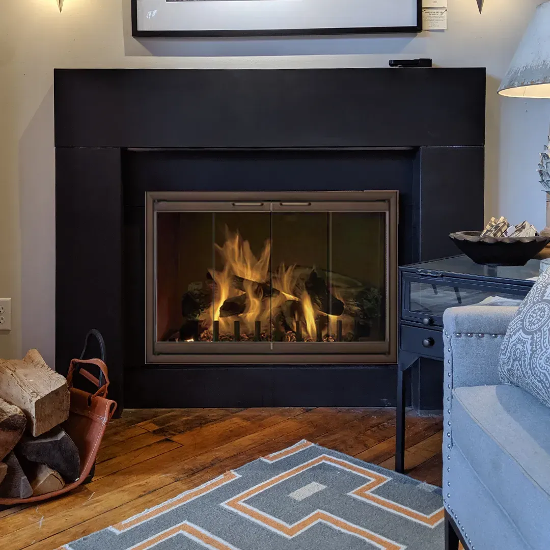 Fireplace with flames, black surround, firewood basket, blue rug, and wooden floor.