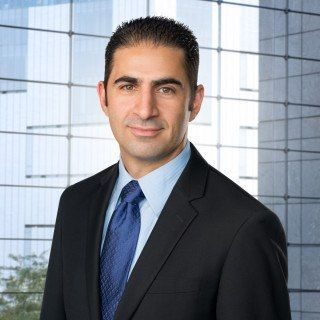 Professional portrait of a man in a dark suit and blue tie in front of an office building