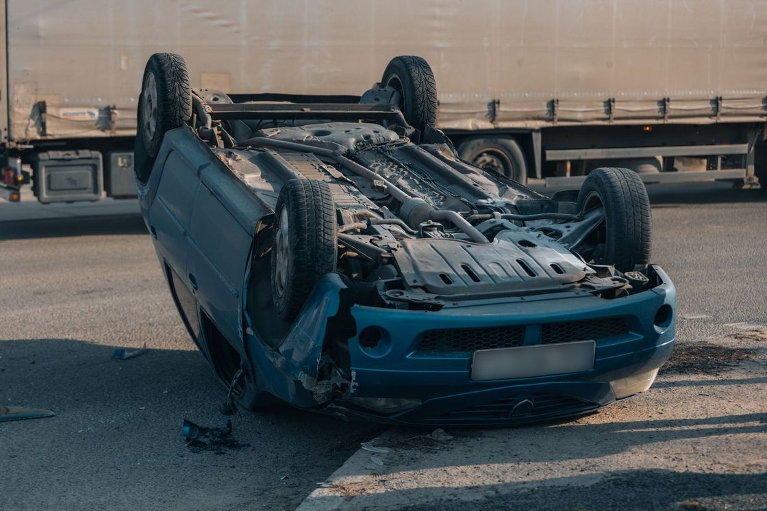 Overturned blue car on highway shoulder, heavily damaged after crash with truck in background