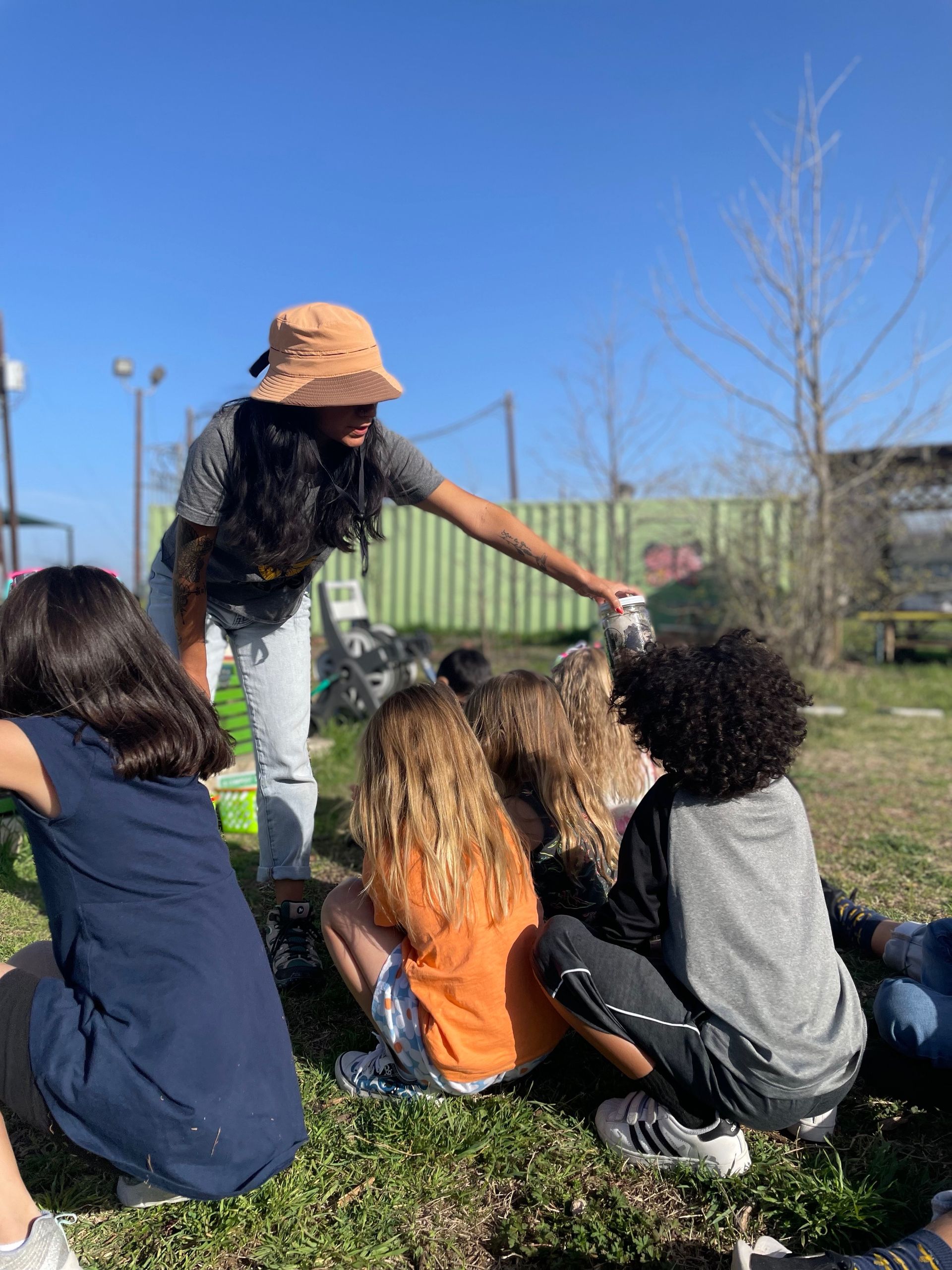 A woman is talking to a group of children sitting on the grass.
