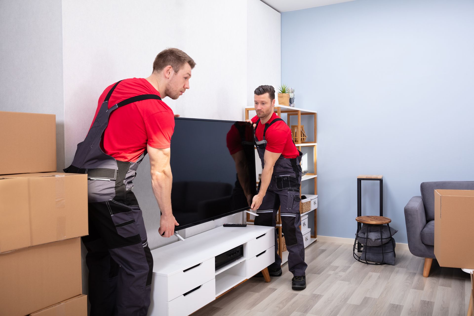Two movers in red shirts and overalls lifting a large TV from a cabinet in a room with packed boxes.