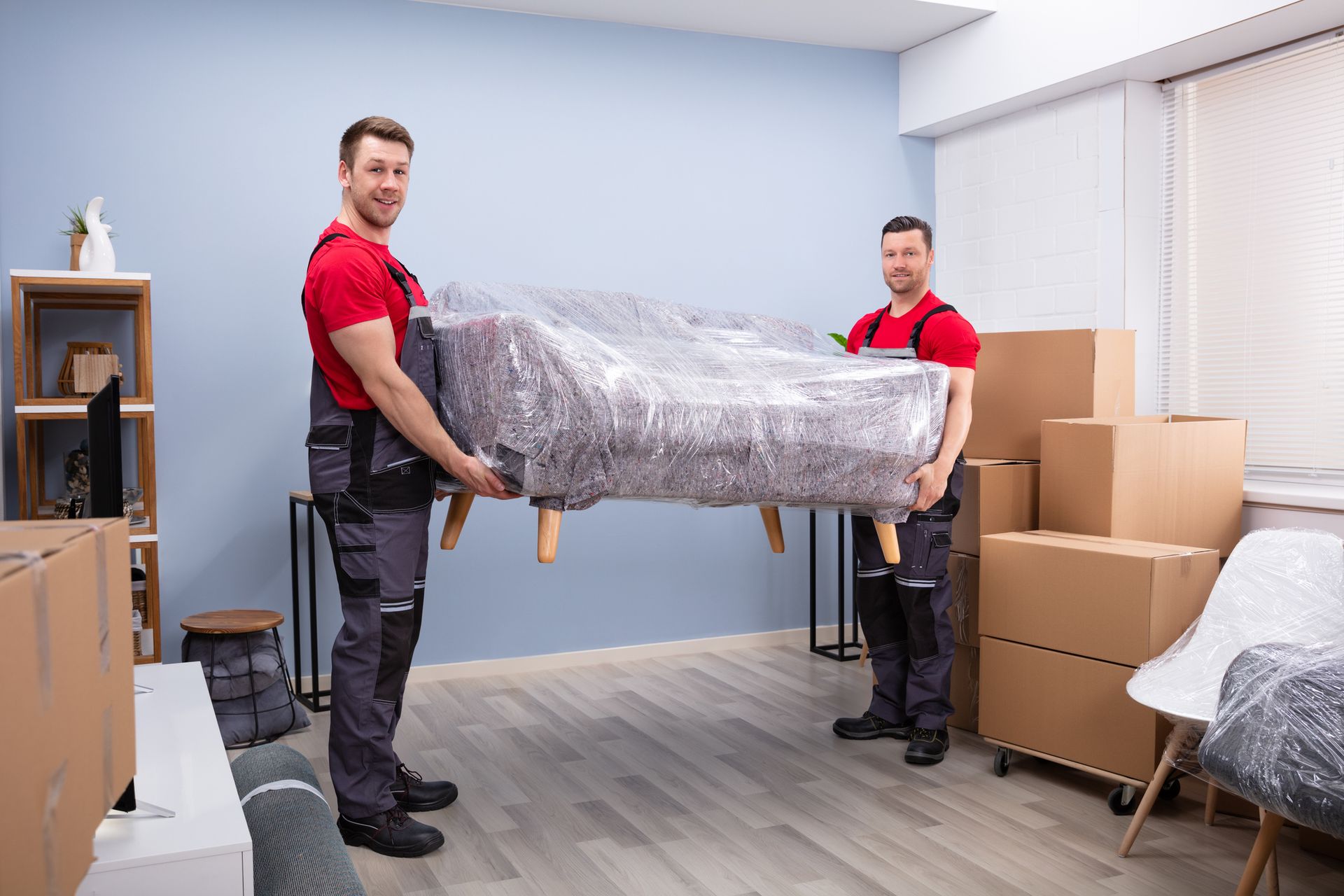 Two male movers, wearing red tees and overalls, hold a sofa covered in plastic wrap, inside a room.