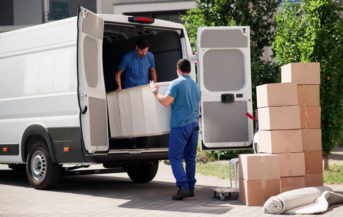 Full-service movers, wearing blue uniforms, are unloading furniture and boxes from a white truck.