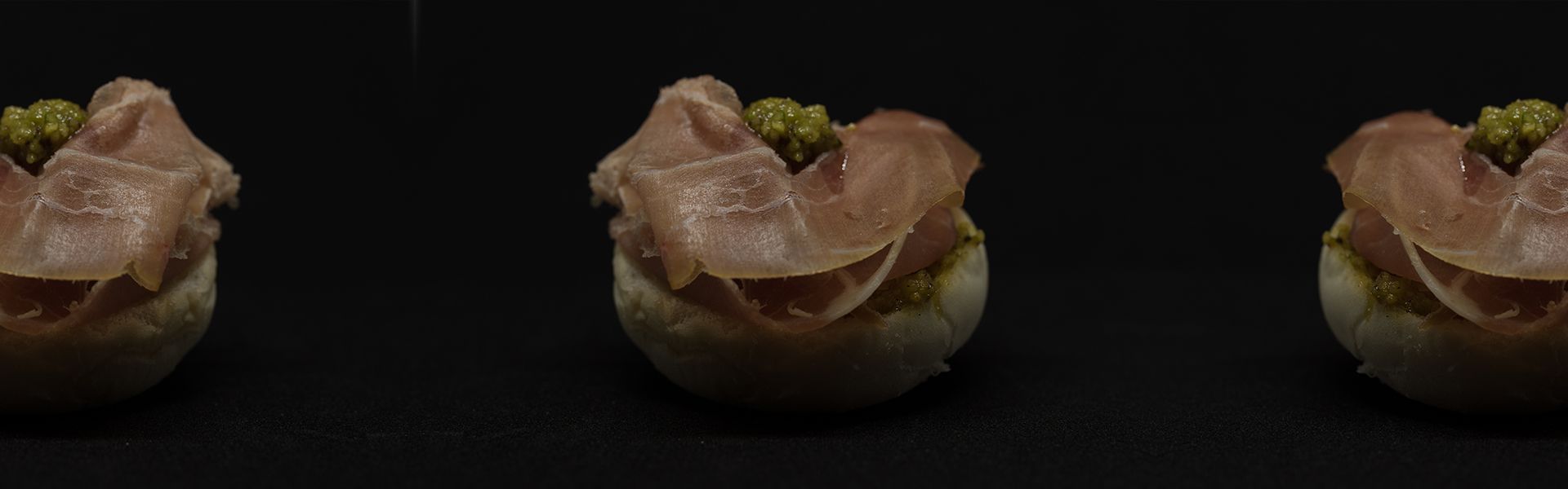 Tre antipasti su una superficie nera; ognuno è composto da carne e guarnizione verde su una base di pane.