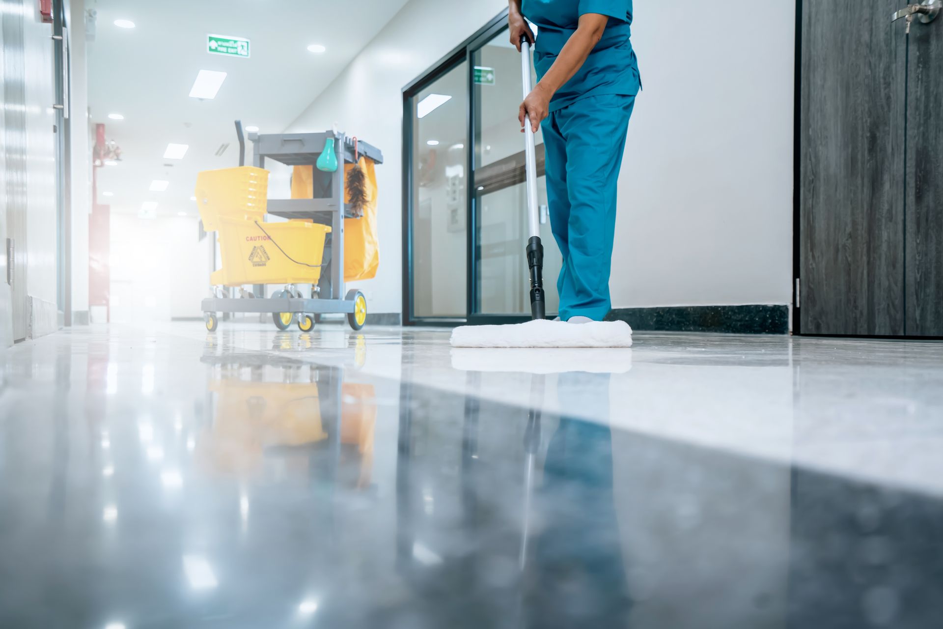 Janitor mopping a shiny hallway floor beside a yellow cleaning cart in a modern building