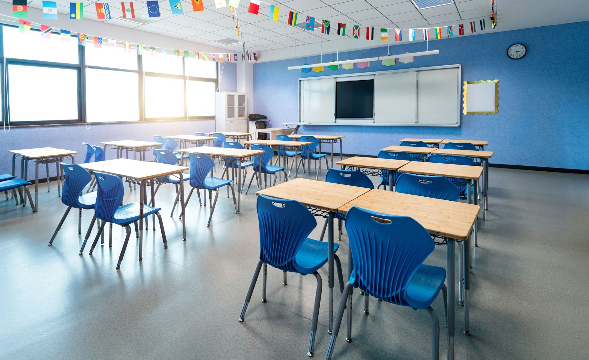 Empty classroom with blue chairs, wooden desks, whiteboard, and bright windows decorated with colorful flags