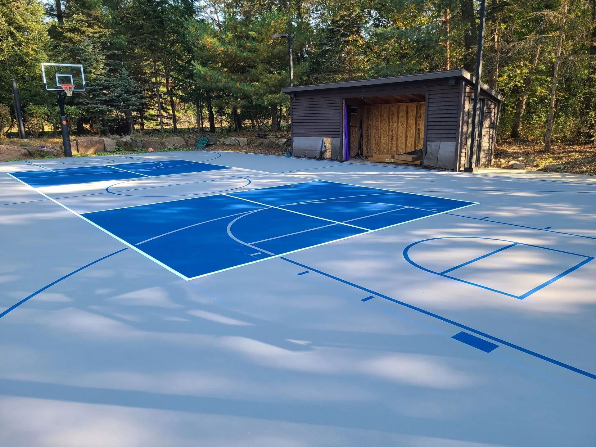 Outdoor basketball court with blue and white painted lines, a hoop, and a small shed at the edge of the court