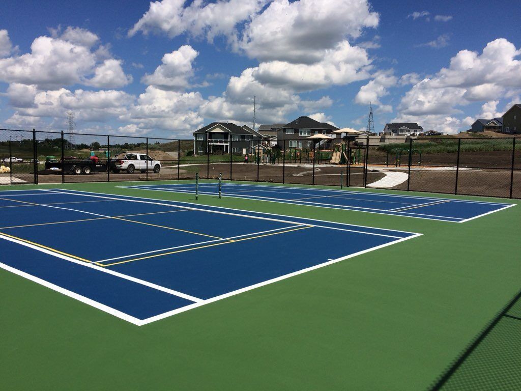 Blue and green tennis courts, black fences, houses in the background, under a bright blue sky with clouds.