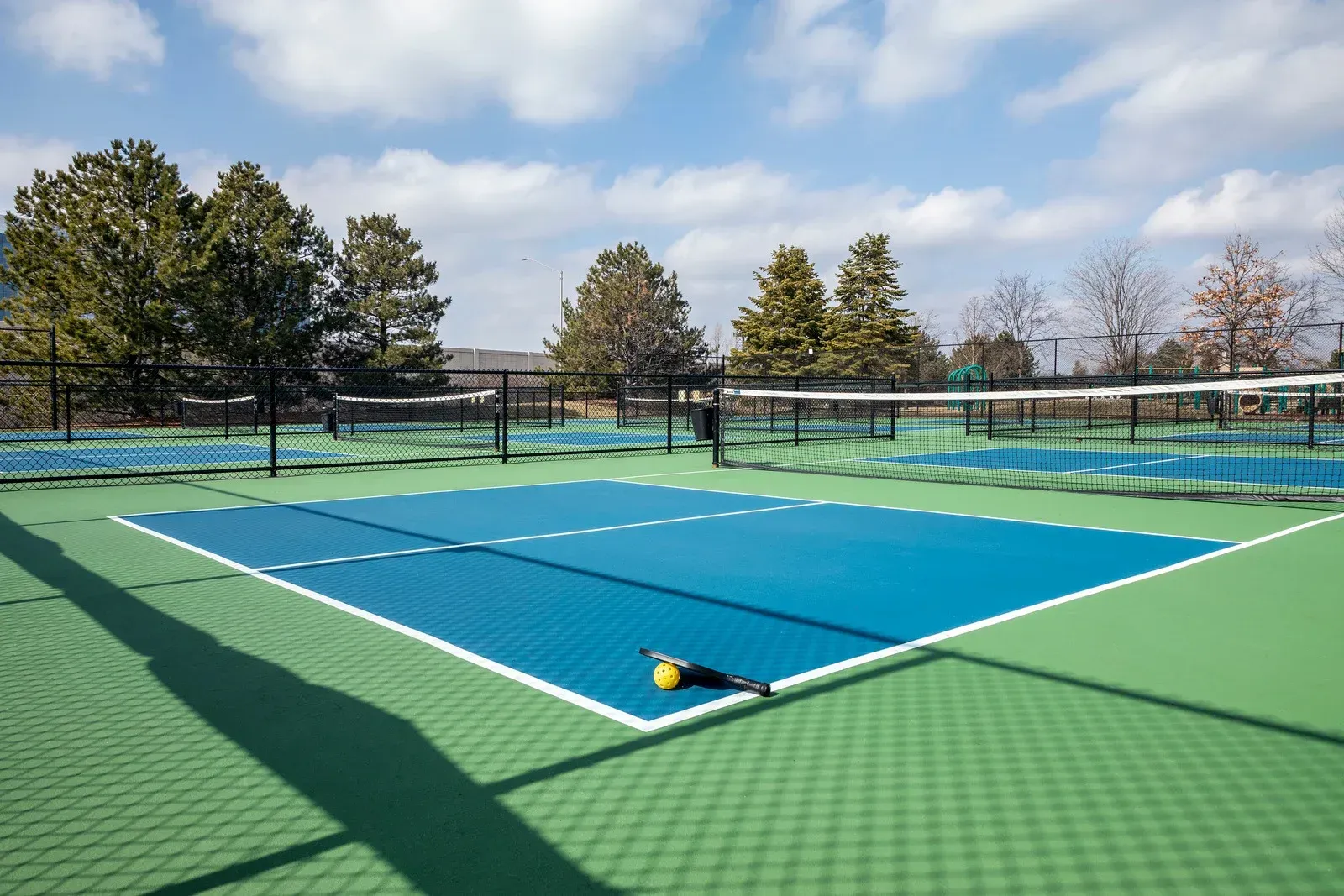 Pickleball court with a blue playing surface, net, and a ball/paddle on the court. Green surround.