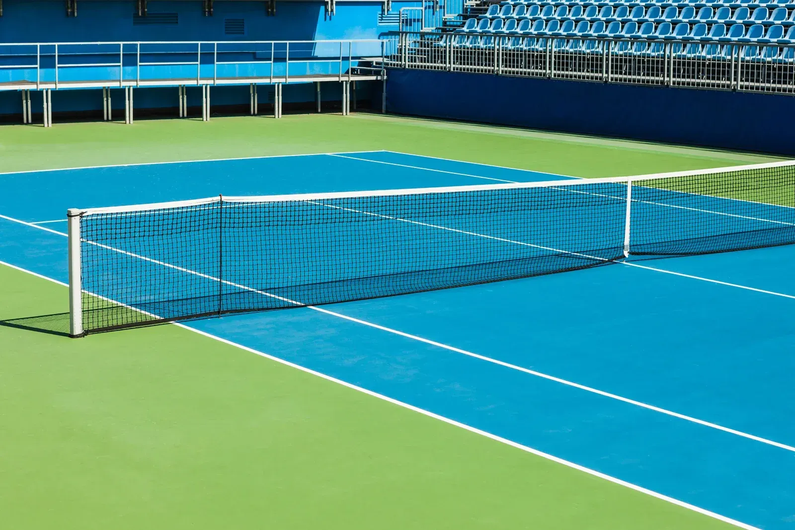 Blue and green tennis court with net, bleachers in the background.