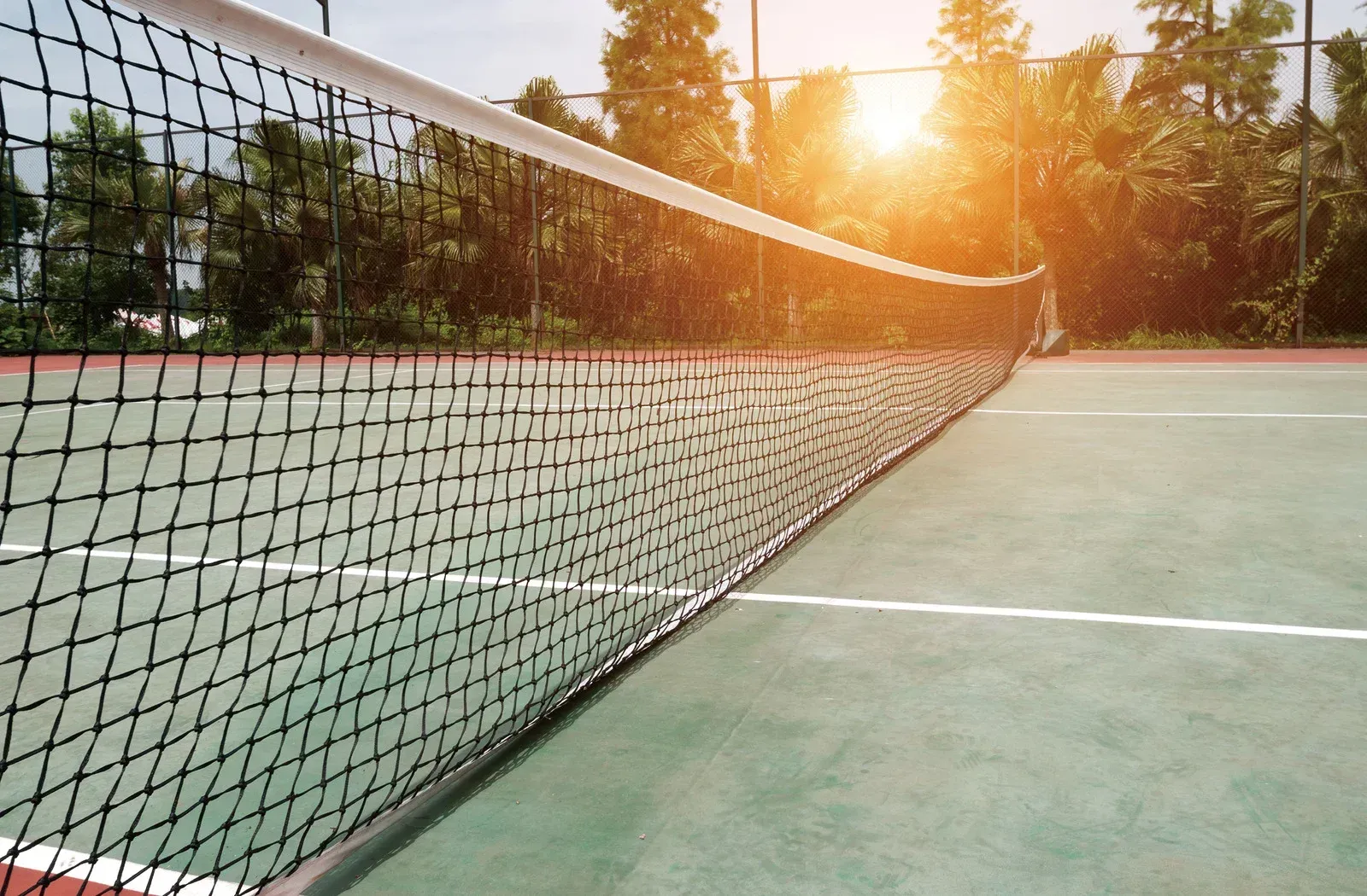 A tennis net on a tennis court with the sun shining through the trees.