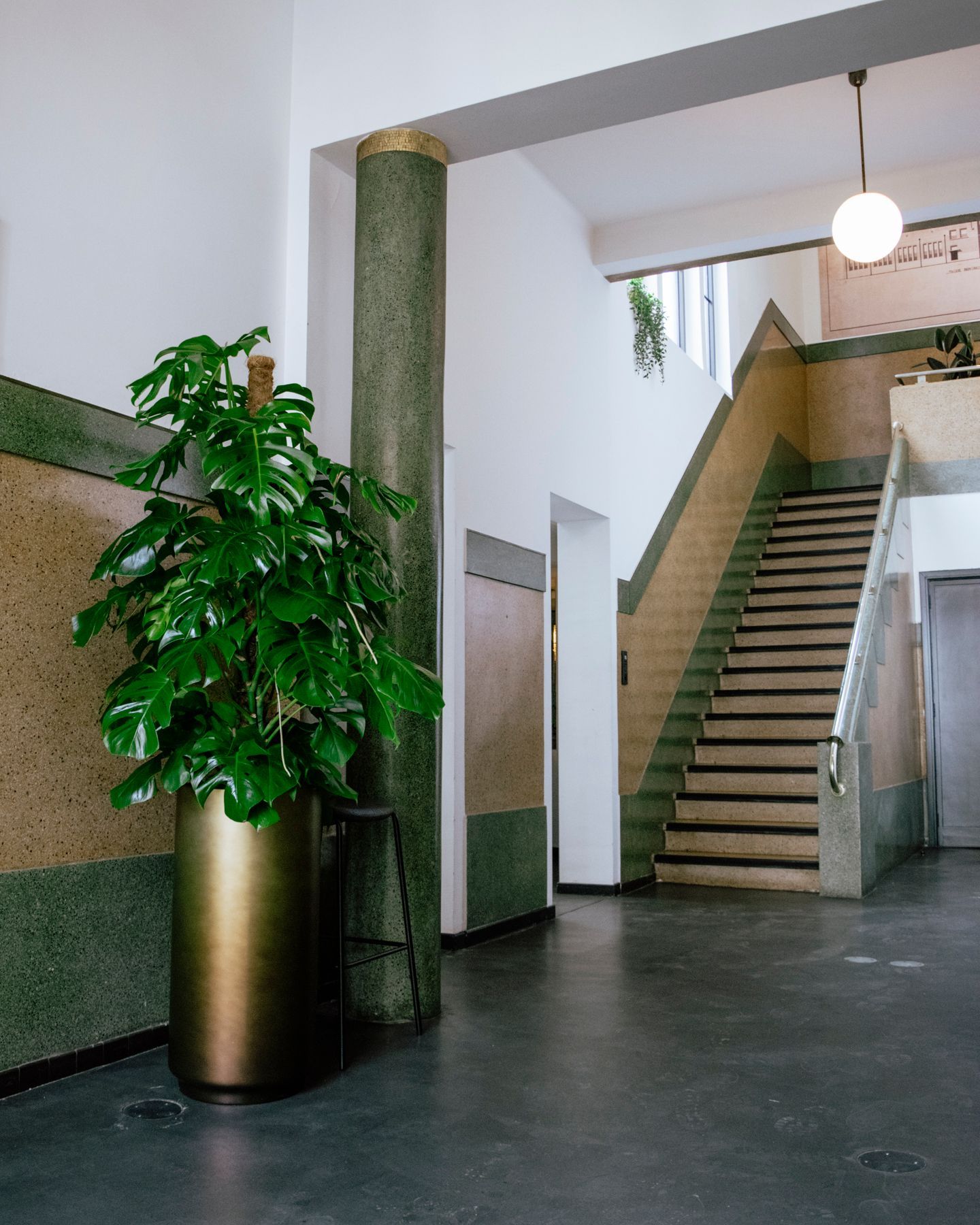 Een grote monstera-plant in een pot staat naast een groene pilaar in de lobby van een modern gebouw met een trap en betegelde muren.
